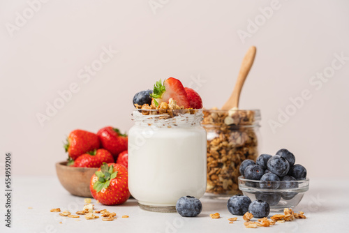 Natural yogurt in jar with granola and fresh strawberries and blueberries on light table and beige background. Healthy homemade breakfast concept