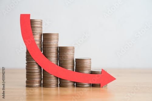 Stack of coins bar chart and red curve graph trending downwards with white wall background on wooden table copy space. Economy recession crisis, inflation, stagflation, business and financial loss.