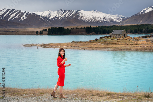 Asian female tourist pose at turquoise color Lake Tekapo with snow capped New Zealand southern alps and Church of Good Shepherd at the background. 