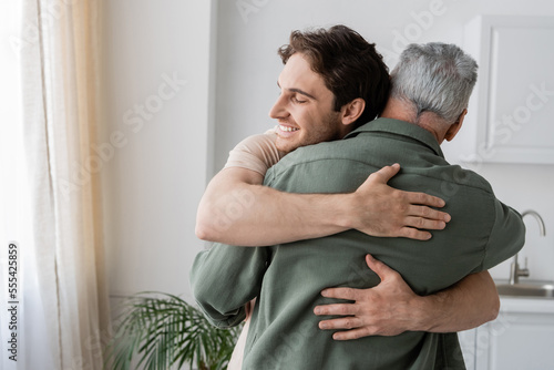 happy young man with closed eyes embracing mature dad in kitchen