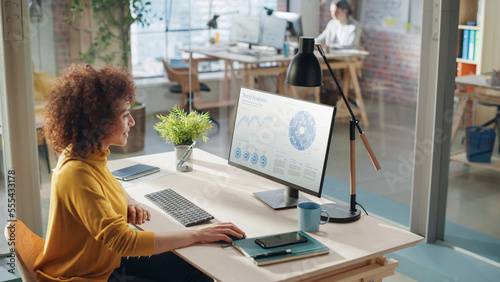 Portrait of a Happy Middle Eastern Manager Working in Creative Office. Business-Driven Stylish Female with Curly Hair Using Desktop Computer Showing Charts and Statistics about Sales on Display.