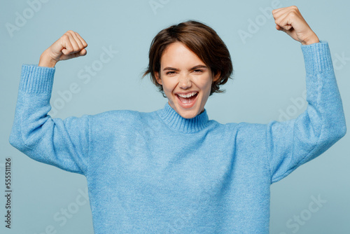 Young strong sporty fitness fun caucasian woman wear knitted sweater look camera showing biceps muscles on hand demonstrating strength power isolated on plain pastel light blue cyan background studio.