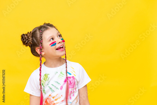 A little girl, painted with paints, looks at your advertisement on a yellow isolated background. The art of painting with paints for children. Art courses for schoolchildren. Copy space.