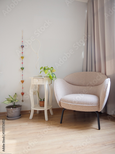 Elegante mueble de lectura beige, acompañado de maceteas con plantas y mesa blanca con lampara y adornos con fondo blanco y cortinas grises