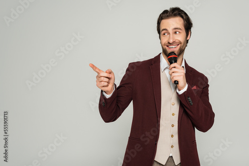 positive showman in formal wear talking in microphone and pointing with finger isolated on grey