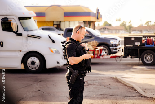 White male caucasian police officer cop standing on street in black uniform, gun on hip, radio walkie talkie talking to dispatch during the day getting information on potential suspect in a bad city.