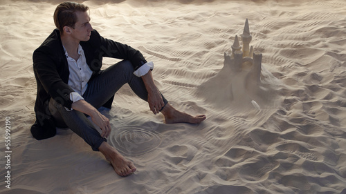 Financier and sand castles, the concept of unprofitable investments. Economic crisis and recession. A young man in a business suit is sitting on the sand next to a sand castle