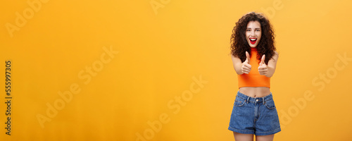 Charismatic ambitious and outgoing charming caucasian woman with curly hairstyle and red lipstick showing thumbs up gesture in like or approval smiling joyfully being supportive over orange wall