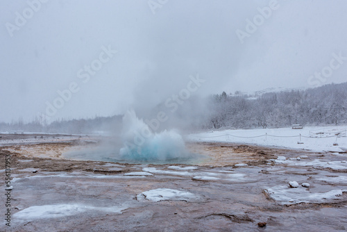 Geyser in eruzione durante una nevicata con cielo grigio e terreno marrone innevato