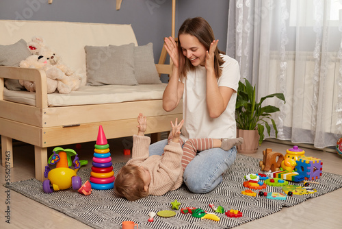 Portrait of smiling happy attractive dark haired woman playing with her infant baby, posing on carpet, having fun with toddler daughter, being photographed among colorful toys.