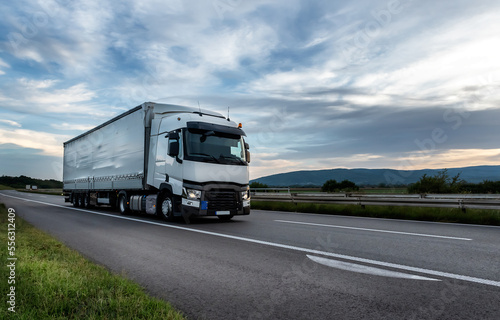 Big White truck driving on the highway through countryside landscape at sunset.