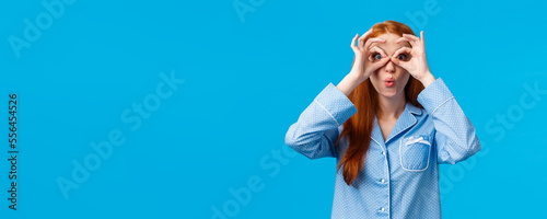Girl saying wow staring startled camera seeing great opportunity for special holidays discount. Attractive redhead female in pyjama looking through fingers and folding lips amused, blue background