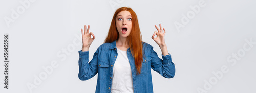 Amused, excited and thrilled good-looking redhead female in denim shirt, open mouth, drop jaw speechless staring with popped eyes camera, showing okay gesture in approval, white background
