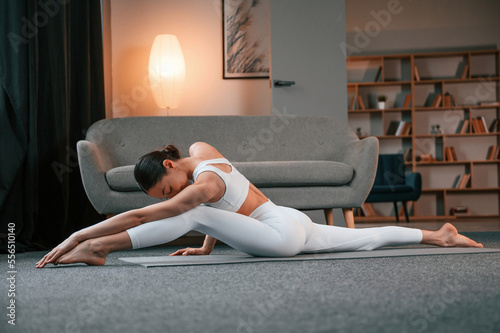 Exercise for legs and spine. Young woman with slim body type in white sportswear is indoors doing yoga