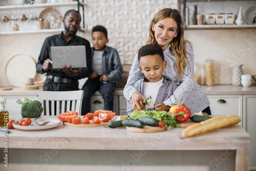 Caucasian young mom and cute small african preschooler son have fun preparing meal in kitchen together, caring father with little boy child on background trying to find recipe of salad using laptop.