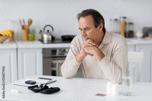pensive and middle aged man with diabetes looking at glucose meter and lancet pen on table