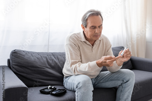 middle aged man taking blood sample with lancet pen while sitting on couch in living room