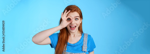 Dreamy curious attractive cheerful redhead woman surprised, look fascinated camera look through fingers okay sign, stand amused, gaze admiration blue background, check out promo