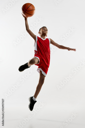 Portrait of boy in red uniform training, playing basketball, throwing ball in a jump over grey studio background. Winning