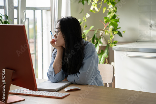 Unmotivated lazy girl sits at home office and looks out window unwillingness to do work. Relaxed Japanese woman sitting at desk with computer procrastinates due to uninteresting and monotonous work 
