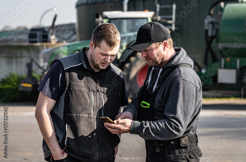  Landwirt erklärt seinen Angestellten technische Details im Hintergrund ist ein Traktor mit Güllefaß sowie Teile einer Biogasanlage zu sehen.