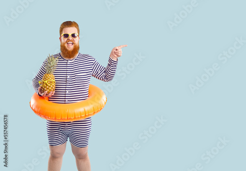 Young redhead happy Caucasian funny fat man tourist with life buoy on belt smiling pointing finger to side holding pineapple dressed in striped beach suit stands. Summer resort concept