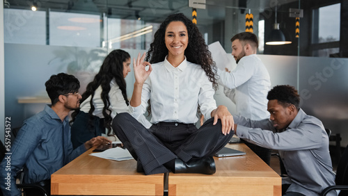 Happy calm smiling businesswoman female leader woman sitting on table in lotus position meditating in office showing ok hand gesture okay sign on background of angry colleagues team throw documents