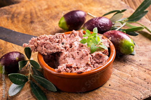 Traditional French goose pate as spread in a bowl with olives served as close-up on a rustic wooden board