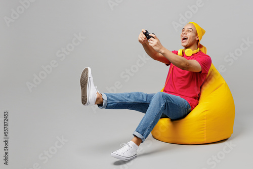 Full body young man of African American ethnicity he wear pink t-shirt yellow hat headphones sit in bag chair hold in hand play pc game with joystick console isolated on plain grey background studio.