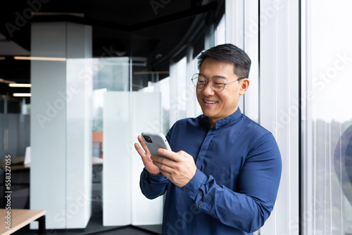 Satisfied with achievement Asian businessman inside office using phone, mature man in shirt near window reading news online from smartphone, successful boss in glasses.