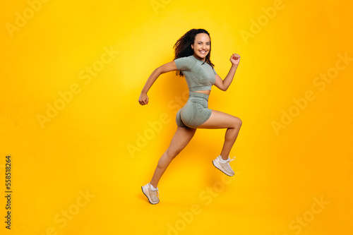 Sports lifestyle. Full length photo of a fitness sporty happy joyful brazilian or latino woman in sportswear, training working out, jumping, running, smiles at camera, isolated orange background