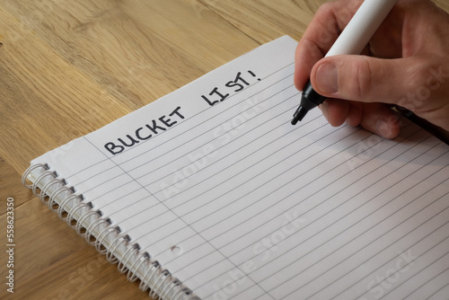 man writing a bucket list on plain lined notepad paper on a wooden desk showing all the things that he wants to do in the rest of his lifetime. The process is intended to succeed in personal ambitions