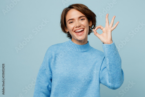 Young satisfied smiling happy fun caucasian woman wear knitted sweater look camera showing okay ok gesture isolated on plain pastel light blue cyan background studio portrait People lifestyle concept