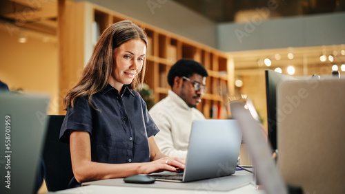 Multiethnic Group of Creative Colleagues Working on Laptops During Day in a Modern Bright Office. A White Woman Smiles Happily After Successfully Finishing a Work Project.