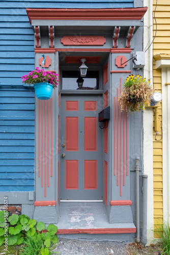 A blue wooden house with an entrance decorated orange and grey in color. There are pots hanging on both sides of the door with purple and yellow flowers. A green plant grows near the door step.