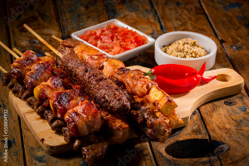 Espetinhos de churrasco em tábua de madeira com acompanhamentos, arroz, vinagrete, farofa e maionese