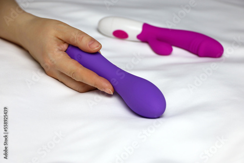 Sex toys in female hand on white sheet. Woman lying on a bed with purple silicone dildo and red vibrator