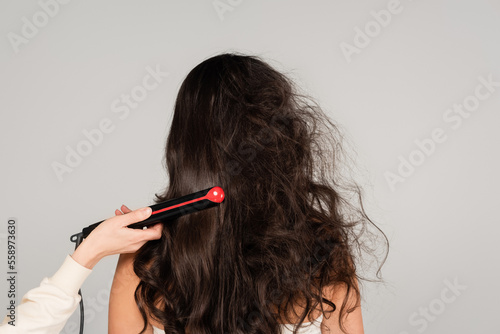 hairdresser straightening hair of curly brunette woman with hair iron isolated on grey.
