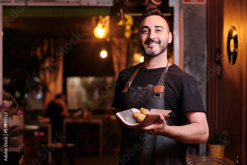 Male waiter in uniform smiling while holding a plate with food in a restaurant.