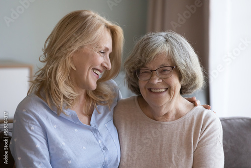 Joyful happy old 80s mom and adult middle aged daughter woman sitting, hugging, on home couch, talking, chatting, laughing, enjoying meeting, leisure time, family relationships