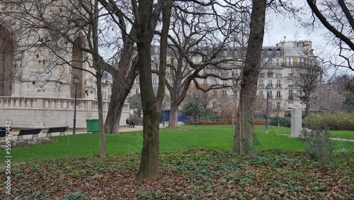 Un parc dans une zone urbaine, avec des bâtiments au fond, un gazon bien entretenu, beaucoup de vert, des arbres sans feuilles, du jardinage, avec des bancs peu occupés et peu de passants, ciel gris e