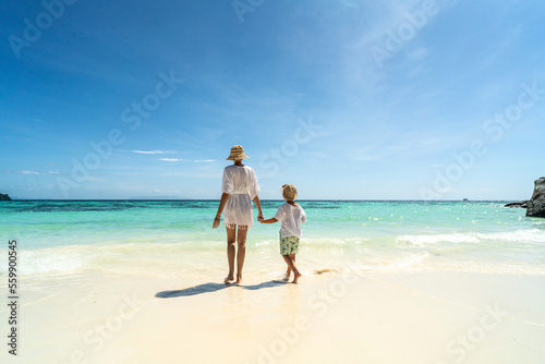 Summer photo of mother with son walking on the sandy tropical beach, relaxing, enjoying sunny day on vacation. Back view. Copy space. Tourism.