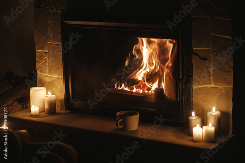 Warm cup of tea and candles at cozy burning fireplace in dark evening room. Fireplace heating in house, electricity blackout. Atmospheric time at fireside in home