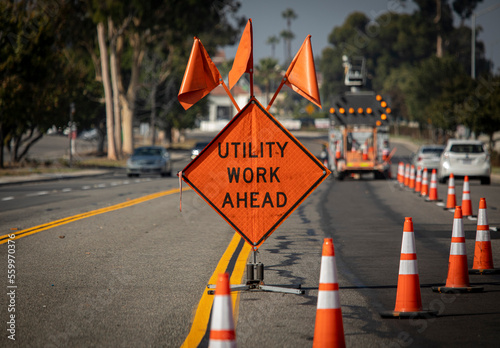 Traffic sign with flags reading Utilitary Work Ahead with traffic cones on road with electronic arrow pointing to the right to divert traffic