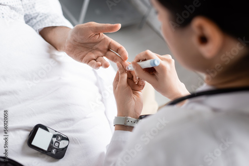 Blurred doctor holding lancet pen near hand of patient with diabetes in hospital ward.