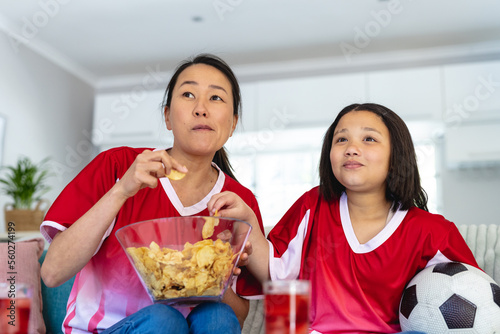 Asian mother and daughter in football shirts watching soccer game, supporting and eating crisps