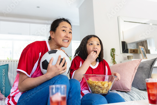 Asian mother and daughter in football shirts watching soccer game, supporting and eating crisps