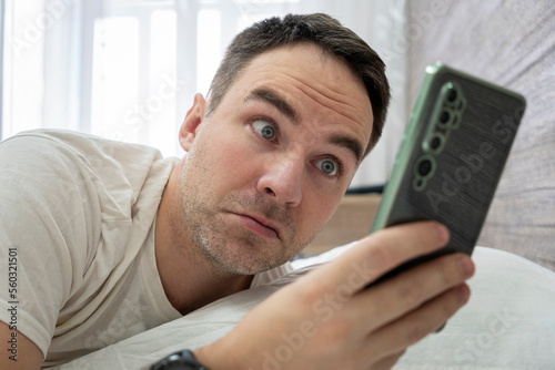 Wake up shocked guy late for work in morning. Man surprised to hear ringing of alarm smart phone, lies in white bed. Surprise from the message on smarfton