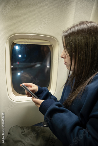 Young woman sitting in an airplane while looking out of the window and using her smartphone - travel concept
