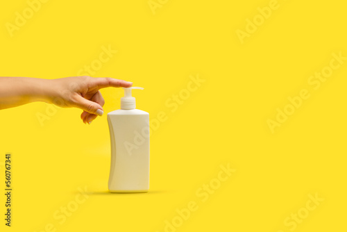 Mano de mujer apretando un bote dispensador de plástico blanco sobre un fondo amarillo brillante liso y asilado. Vista de frente y de cerca. Copy space